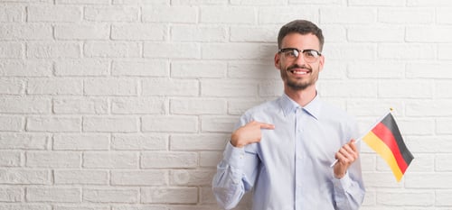 Preview: Young Adult Man Over Brick Wall Holding Flag Of Germany With Surprise Face Pointing Finger To