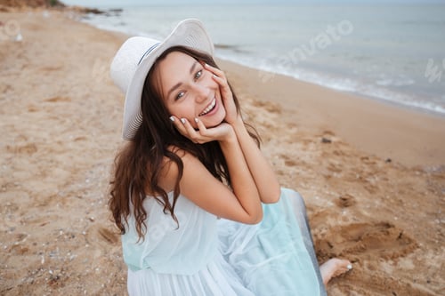 Preview: Cheerful Lovely Young Woman In White Hat And Dress Sitting And Smiling On The Beach