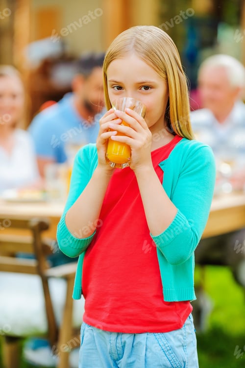 Preview: Enjoying Fresh Juice. Cute Little Girl Drinking Orange Juice And Smiling While Her Family Sitting