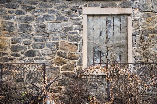 Preview: Abandoned Stone Building With Cemented Barred Window And Fence With Overgrown Weeds