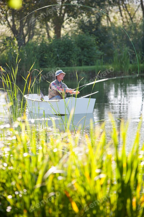 Preview: Vertical Wide Shot Of An Elderly Man Fishing In A Rowboat Over A Lake With Tall Grass Blades In The