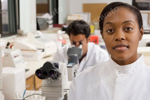 Preview: Female Doctor Looking At The Camera With A Male Doctor Looking Through A Microscope In The