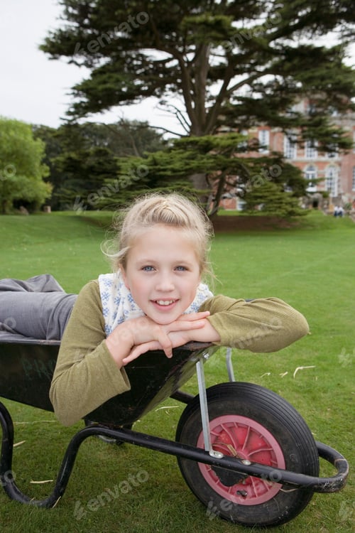 Preview: Side View Of A Girl Lying In Wheelbarrow On Grass In Large Country Garden Looking At The Camera.