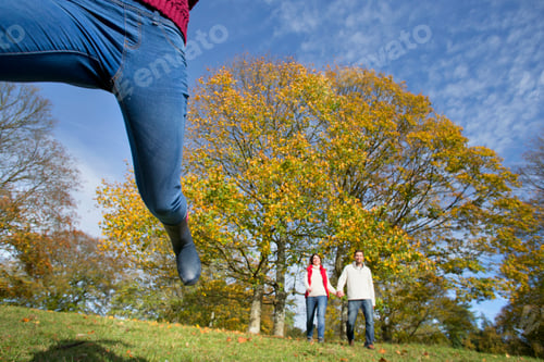 Preview: Child Leaping In An Autumnal Forest With Her Parents Holding Hands And Walking In The Background.