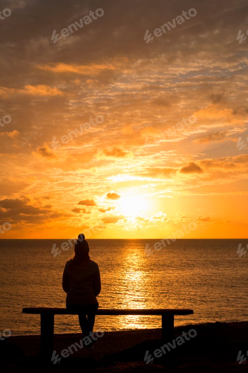 Preview: Vertical Shot Of The Silhouette Of A Woman Sitting On A Bench Looking At The Tranquil Sunset Over