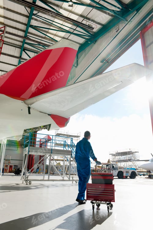 Preview: Engineer Rolling A Cart Loaded With Toolboxes Below The Tail Of A Passenger Jet At A Hangar.