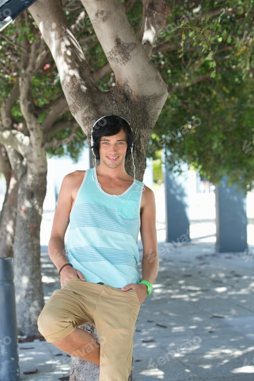 Preview: Vertical Portrait Of A Smiling Young Man Wearing Headphones And Leaning Against A Tree In A Park