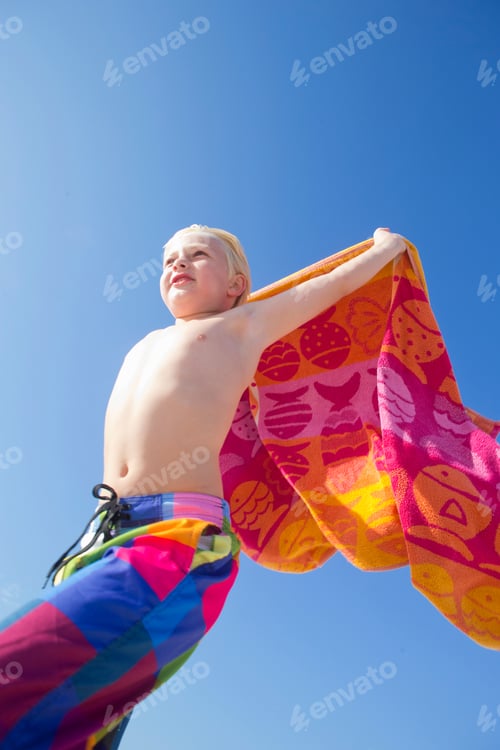 Preview: Vertical Shot Of A Boy Holding A Towel Above His Head And Running On A Sunny Beach.