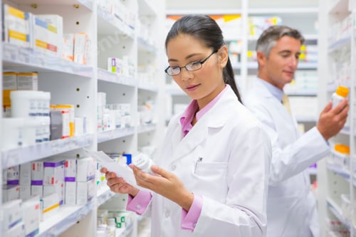 Preview: Female Pharmacist Reading The Label On A Medicine Pot At A Pharmacy With A Colleague In The