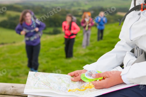 Preview: Close-Up Shot Of A Girl Holding A Compass And Map In A Green Field With Group On Friends In The