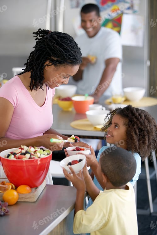 Preview: Mother Serving Fruit Salad To Children With Father In Background At Home.