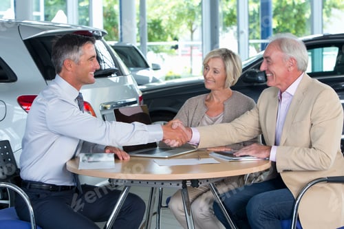Preview: Smiling Salesman Shaking Hands With Couple At A Table In A Car Dealership Showroom