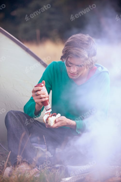 Preview: A Young Man Sitting Next To A Tent, Putting Tomato Ketchup On A Hot Dog