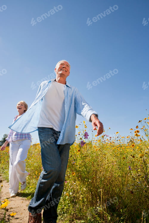 Preview: Vertical Shot Of An Enthusiastic Elderly Couple Holding Hands And Running Along A Path In A Meadow