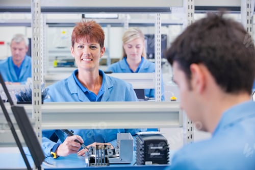 Preview: Female Technician In An Apron Smiling At The Camera While Assembling Circuit Boards With Coworkers