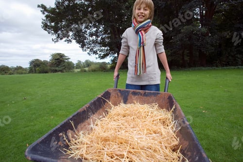 Preview: A Blond Boy Wearing Colorful Scarf Holds A Wheelbarrow Full Of Hay On A Lawn.
