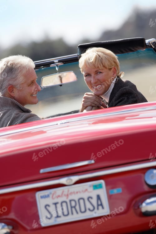 Preview: Senior Couple Driving Classic Red Convertible Car On Road Trip At Camera