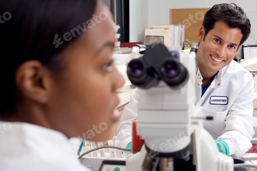 Preview: Male Doctor Smiling At The Camera With A Female And A Microscope In The Foreground.