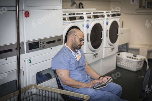 Preview: Mature Man In Surgical Scrubs Using Touchscreen On Digital Tablet In Laundry Room