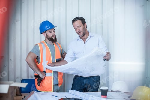 Preview: Construction Foreman Looking Over Blueprint With Worker At Desk In Portable Cabin