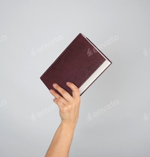 Preview: Hand Holds A Closed Book In Hardcover On A Gray Background