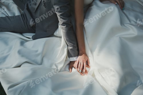 Preview: Elegant Wedding Couple Is Sitting On White Satin Dress. Slim Bride Is Holding Hands With Groom In