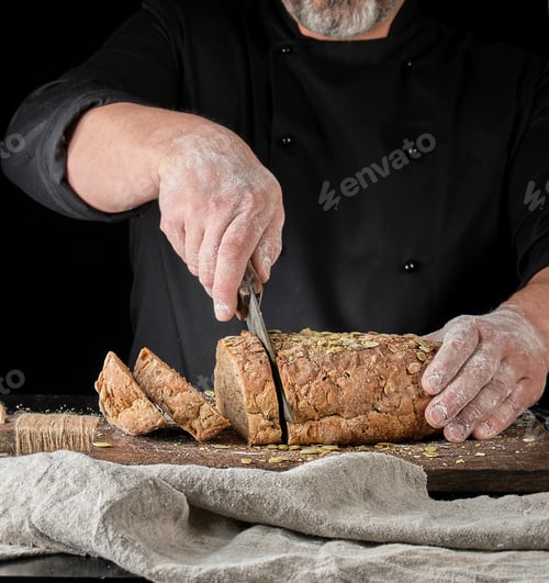 Preview: Baker Cuts A Knife Into Slices Of Rye Bread With Pumpkin Seeds On A Brown Wooden Board, Black