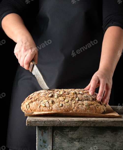 Preview: Female Hands Hold Oval Baked Rye Bread With Pumpkin Seeds, Dark Background