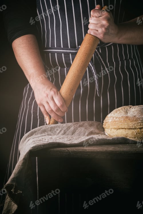 Preview: Woman In An Apron Holds A Wooden Rolling Pin Next To Baked Round Bread On A Wooden Table, Dark