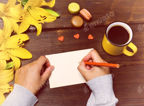 Preview: Female Hands With A Red Pencil Near A White Blank Postcard On A Brown Wooden Table, Next To A Cup