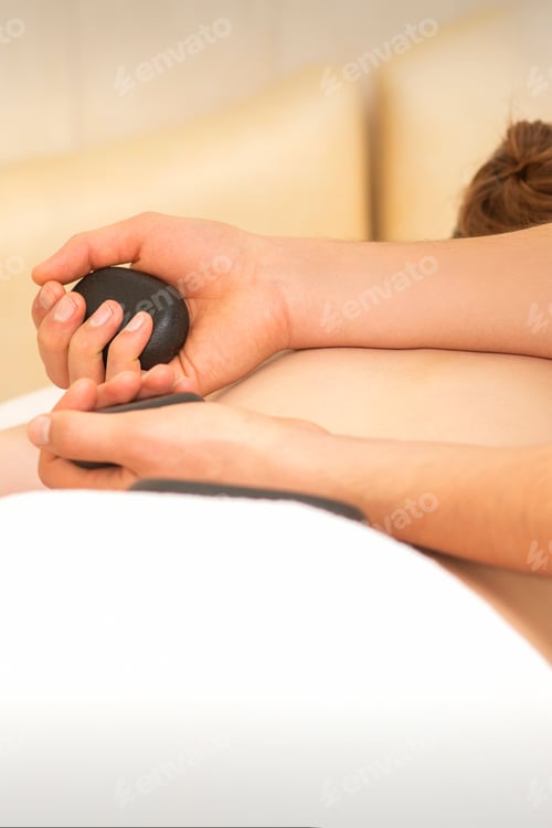 Preview: Hands Of Masseur Hold Black Hot Stones Massaging Back Of A Young Adult Woman In Spa Salon