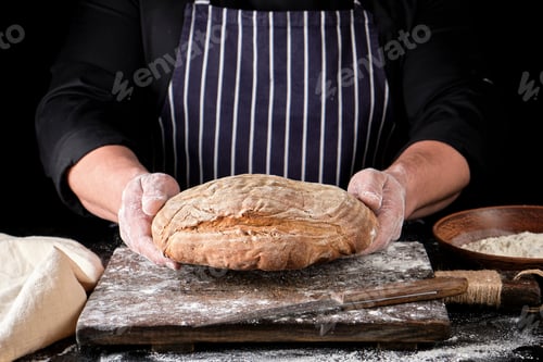 Preview: Male Hands Are Holding Brown Baked Rye Bread Over Wooden Board With Flour, Black Background