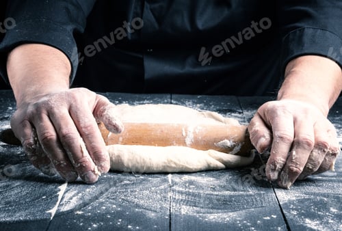Preview: Chef In A Black Tunic Rolls A Dough For A Round Pizza On A Black Table With A Wooden Rolling Pin
