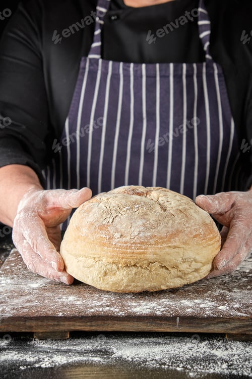Preview: Chef In Black Uniform And Apron Holds Round Baked Bread Made From White Wheat Flour, Close Up