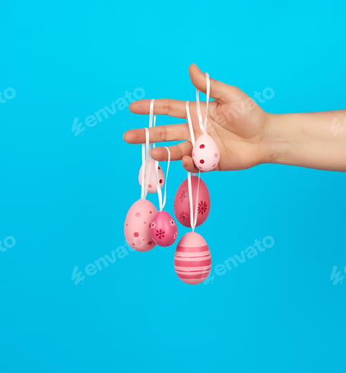 Preview: Female Hand Holds Easter Colored Pink Eggs On A Rope, Blue Background