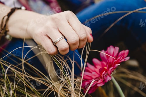Preview: Young Woman With Ring In Her Hand Resting On The Grass Near A Pink Flower.