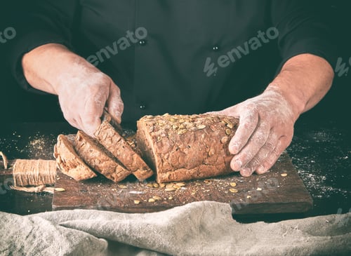 Preview: Baker Cuts A Knife Into Slices Of Rye Bread With Pumpkin Seeds On A Brown Wooden Board, Black