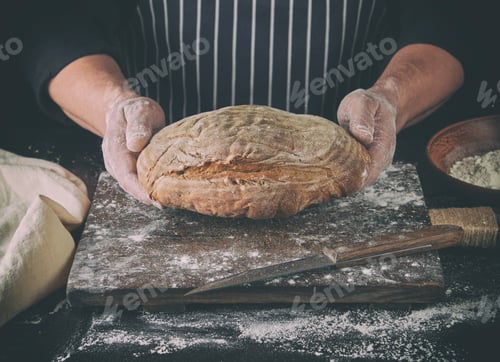 Preview: Male Hands Are Holding Brown Baked Rye Bread Over Wooden Board With Flour, Black Background
