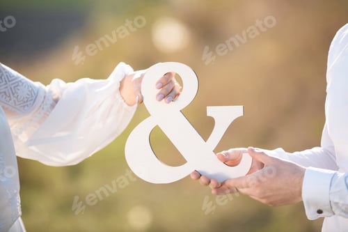 Preview: Bride And Groom Holding White Wooden Sign "And" At Sunset