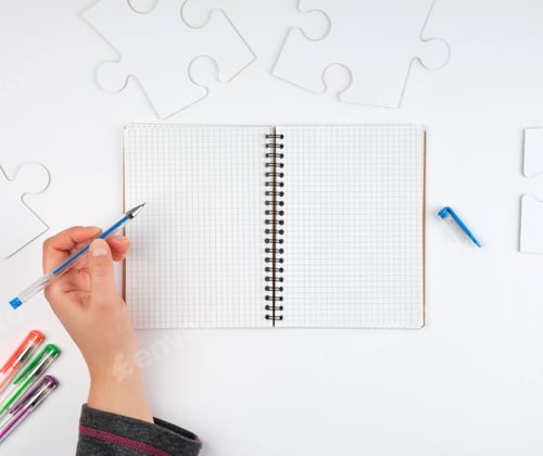 Preview: Open Notebook In A Cell, Hand Holding A Blue Pen, Next To It There Are Big White Puzzles, Top View