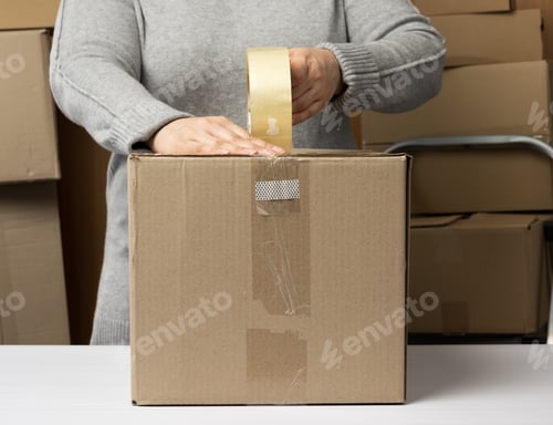 Preview: Woman In A Gray Sweater Holds A Roll Of Duct Tape And Packs Brown Cardboard Boxes On A White Table
