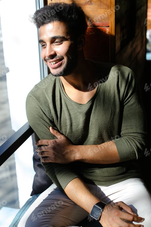 Preview: Man Sitting And Puts Hand On Table. Boy Has Dimples, Beard, Full Lips And Black Hair. Male Dressed