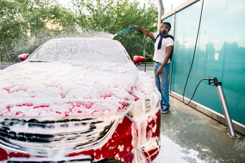 Preview: Young Black Man Cleaning His Red Luxury Car With Foam Spray Outside At Self Car Wash Station