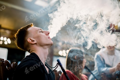 Preview: Caucasian Young Man In Black T-Shirt Smoking A Hookah While Sitting At The Table In Cafe Indoors
