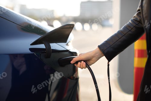 Preview: Cropped View Of Female Hand, Holding Power Cable Supply Plugged In Electric Car, Charging