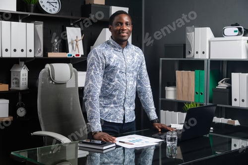 Preview: Portrait Of Three-Quarters Long Young Black Businessman Standing Near Desk In Office