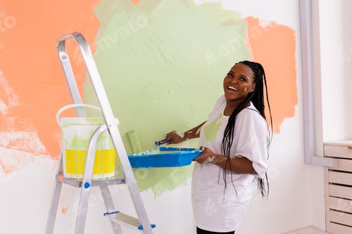Preview: Young Happy African American Woman Painting Interior Wall With Paint Roller In New House. A Woman