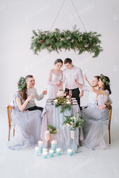 Visualização: Casal de noivos sorrindo na mesa de casamento decorada. Damas de honra e padrinhos bebem champanhe