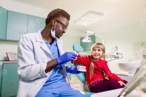 Preview: Little Girl Is Sitting In Dental Chair And Smiling To African American Dentist With Artificial Jaw