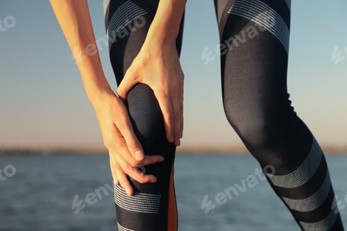 Preview: Young woman in sportswear having knee problems near river at sunset, closeup